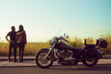 A couple is standing in a field and looking at the sky into the distance. The couple arrived on a motorcycle. The guy and the girl took a break, date, extreme. Couple near the bike. Traveling