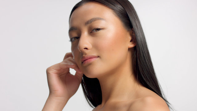 mixed race asian model in studio beauty shoot Model poses to a camera, straight hair Ideal skni and no makeup makeup Head and shoulders crop touches her skin with hand
