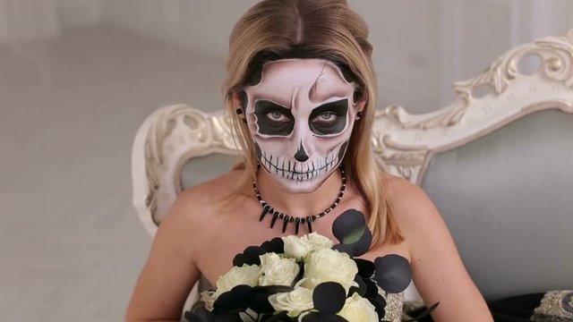 Creepy zombie woman with makeup in the shape of a skeleton sitting on a vintage sofa with a bouquet of black flowers. Portrait of woman with terrifying halloween skeleton makeup. Black wedding.
