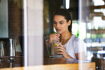 Beautiful woman is thinking about something good, while is waiting file downloads on her laptop computer. Pretty female student is enjoying juice, while is resting after video conference via net-book