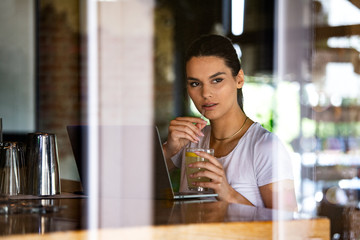 Beautiful woman is thinking about something good, while is waiting file downloads on her laptop computer. Pretty female student is enjoying juice, while is resting after video conference via net-book