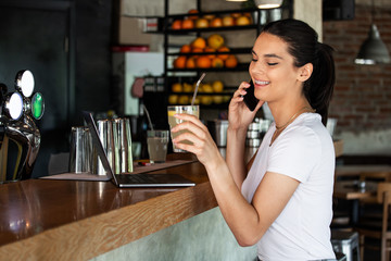 Young charming woman calling with cell telephone while sitting alone in juice bar during free time, attractive female with cute smile having talking conversation with mobile phone while rest in bar
