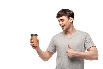 smiling handsome man looking at disposable cup and showing thumb up isolated on white