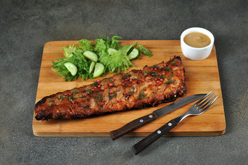 Pork ribs barbecue on a wooden board. Lettuce with cucumbers, mustard and cutlery. Close-up.