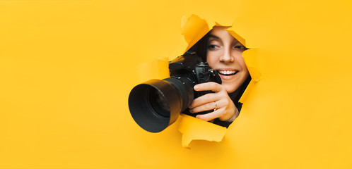A young paparazzi girl holds a reflex camera and looks through a torn hole in yellow paper. The concept of smile and laughter from what he saw. Copy space. - obrazy, fototapety, plakaty