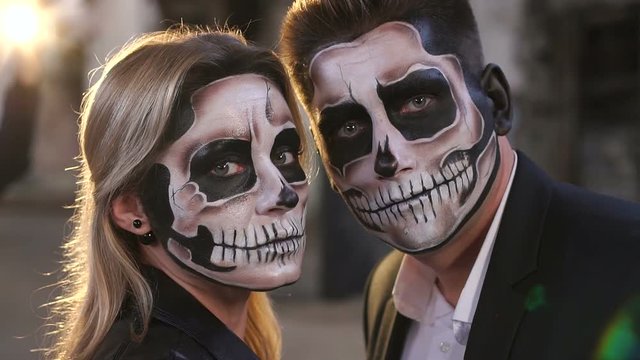 Couple in wedding clothes and with a terrible makeup for Halloween stand against a burning fire in the dark among the smoke. Halloween loving couple with skull make-up. Halloween or horror theme.