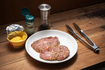 marinade raw pork steak in white plate beside with pepper arigano  cheese and tongs on wooden table