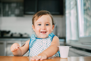 Cute, funny girl eating yogurt during the day in the kitchen in a blue dress