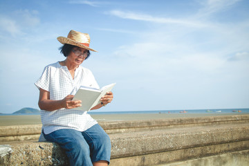 An elderly Asian woman reading a book at the concrete bridge by the sea to relax and breathe fresh...