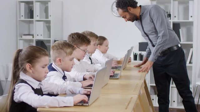 Medium shot of 9-year-old elementary schoolchildren working on laptops at ICT class, and charismatic black male teacher walking from one child to another, checking and giving advice and instructions