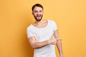 Happy excited bearded man in stylish white T-shirt pointing away at copy space with finger isolated over yellow background, direction, this way. look here, place for advert. copy space