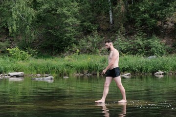 A guy with a Caucasian-type beard walks along the surface of the water in a river or a lake in a forest. The concept of human unity with nature, travel and lifestyle. Copy space.
