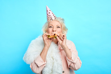 happy mature childish careless woman in party hat making a noise in the party. isolated blue background, studio shot. celebration, happiness . lifestyle - 278581019