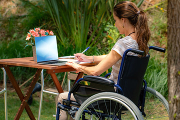 Side view of young handicapped woman in wheelchair working on laptop in garden.