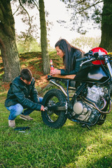 Fototapeta premium Young man fixing his damaged motorcycle while his girlfriend looks at him taking a beer outdoors