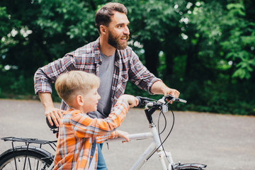 Obraz premium Side view of handsome dad and his cute little son walking with bike in green summer park, looking forward and smiling.