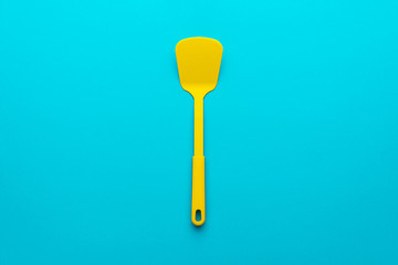 Top view of vivid plastic kitchen utensil. Minimalistic flat lay image of yellow spatula over turquoise blue background with copy space. Central composition of turning spatula on blue table.