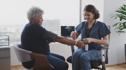 Nurse Wearing Scrubs In Hospital Office Checking Senior Female Patients Blood Pressure