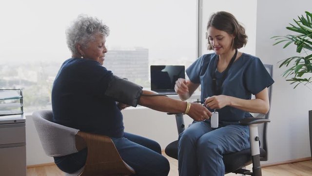 Nurse Wearing Scrubs In Hospital Office Checking Senior Female Patients Blood Pressure