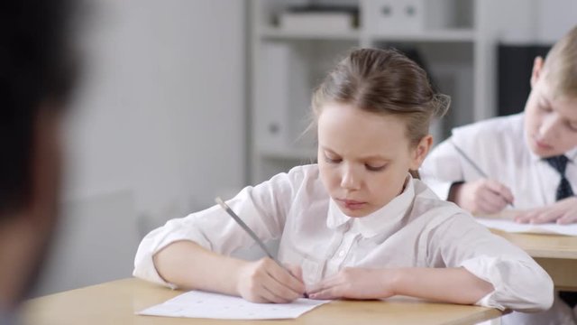 Chest-up shot of frowning 9-year-old Caucasian schoolgirl, wearing uniform white blouse, sitting at desk and filling out test answer sheet, classmates sitting around, and teacher in blurred foreground
