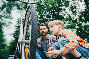 An adult hipster beard father and his elementary age little son wearing in plaid shirt repairing bicycle together outside on a sunny summer day in park.