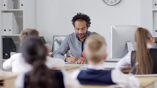 Waist-up shot of smiling African American male teacher sitting at his desk in front of classroom, asking questions to schoolchildren and putting marks and notes on lesson report sheet