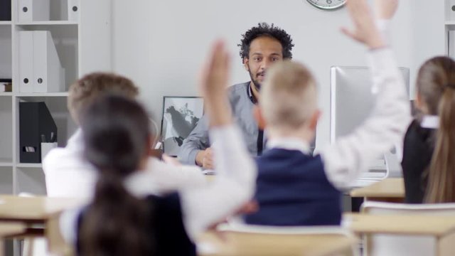 Medium shot of happy young Afro-American male teacher asking questions at lesson in elementary school, pupils raising hands, teacher pointing to boy, and then continuing to explain new material