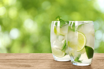 Glasses of refreshing drink with lime slices and mint on wooden table against blurred background, space for text