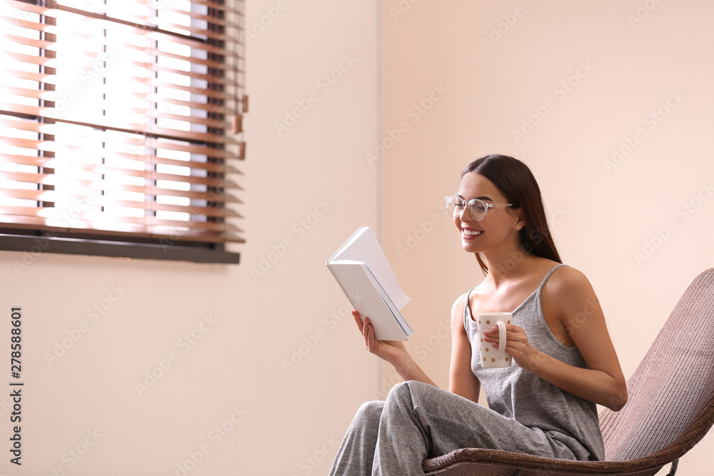 Poster Young woman reading book near window with blinds at home. Space for text