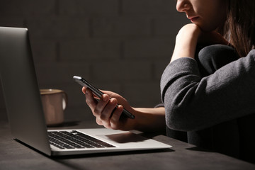 Woman using smartphone at table with laptop in dark room, closeup. Loneliness concept