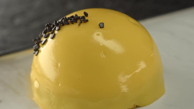Coconut mousse dessert with mango and passion fruit, yellow mirror glaze coating and chocolate decor. Chocolate decoration falls on dessert. Modern mousse cake covered with yellow glaze on black plate