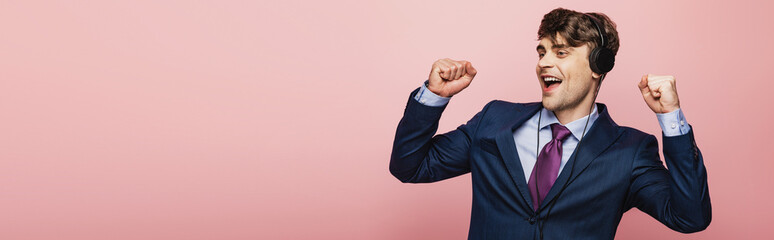 panoramic shot of excited businessman dancing while listening music in earphones on pink background