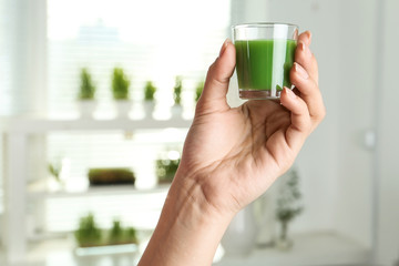 Woman holding shot glass of wheat grass juice indoors, closeup