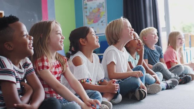 Group Of Laughing Elementary School Pupils Sitting On Floor Listening To Teacher 