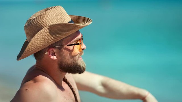 Handsome European guy in sunglasses and hat relaxing on beach at seascape background