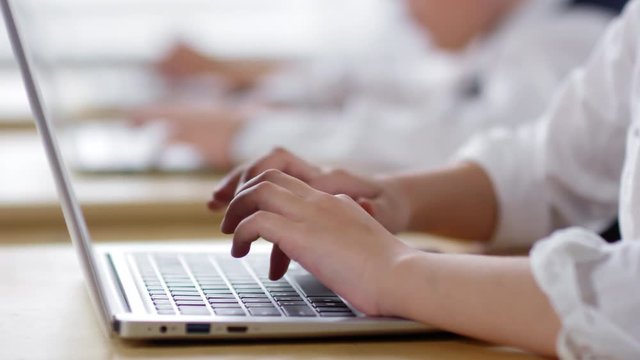 Close-up hands shot of young schoolchild, dressed in white shirt with rolled-up sleeves, sitting at desk in classroom and typing on laptop, with classmates in blurred background
