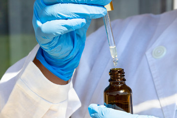 Woman hand in white coat and blue medical gloves holding pipette dropper with drop of natural oil...