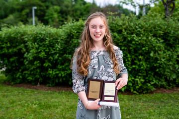 Young teen girl/Middle school student standing in front of a bush/garden after her graduation ceremony while holding a diploma and academic awards.
