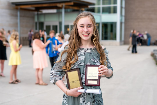 Young Teen Girl/Middle School student standing in front of school with awards and diploma after Grade 8/Middle school graduation ceremony with blurred people in background.