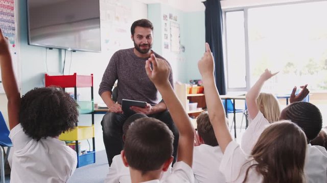 Male Teacher Holding Digital Tablet Teaches Group Of Uniformed Elementary Pupils In School Classroom