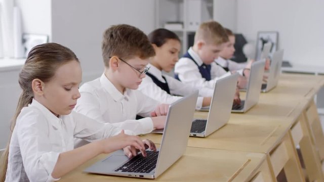 Medium shot of five Caucasian primary schoolchildren, wearing school uniform, sitting in row at their desks and diligently working on laptops during computer education class