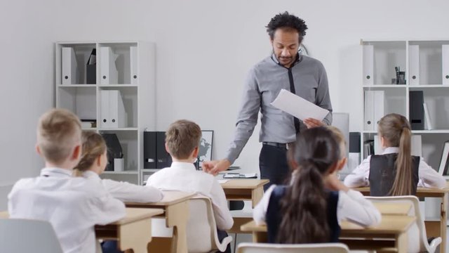 Medium full shot of enthusiastic African American male teacher walking between rows of desks during lesson and distributing answer sheets to preteen schoolchildren for module test