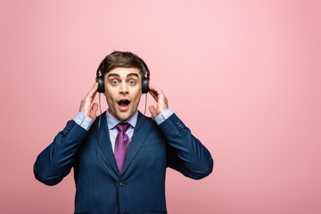surprised businessman looking at camera while listening music in earphones isolated on pink