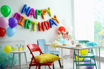 Table with treats and phrase HAPPY BIRTHDAY made of colorful balloon letters in living room