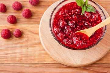 Glass small saucer with raspberry jam with fresh raspberries and mint close-up on wooden background.Copy space