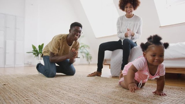 Baby Girl Learning To Crawl At Home As Parents Sit On Floor And Give Encouragement