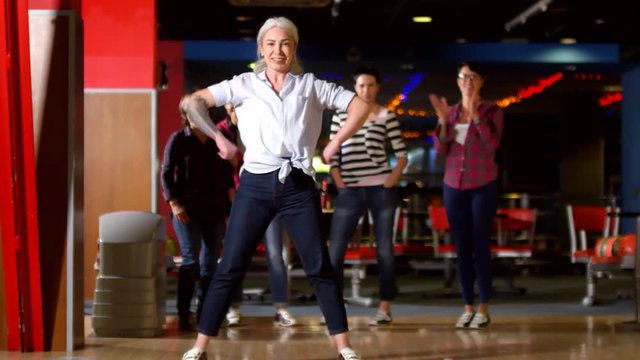 Medium shot of happy mature woman throwing bowling ball and celebrating knocking over pins with group of her female friends