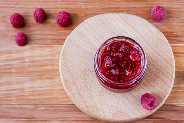 Glass small jar with raspberry jam with fresh raspberries close up on wooden background.Copy space