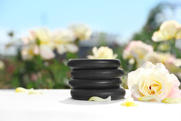 Stack of spa stones and beautiful rose on white table in blooming garden, space for text. Harmony and zen