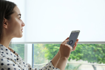 Woman using smart home application on phone to control window blinds indoors